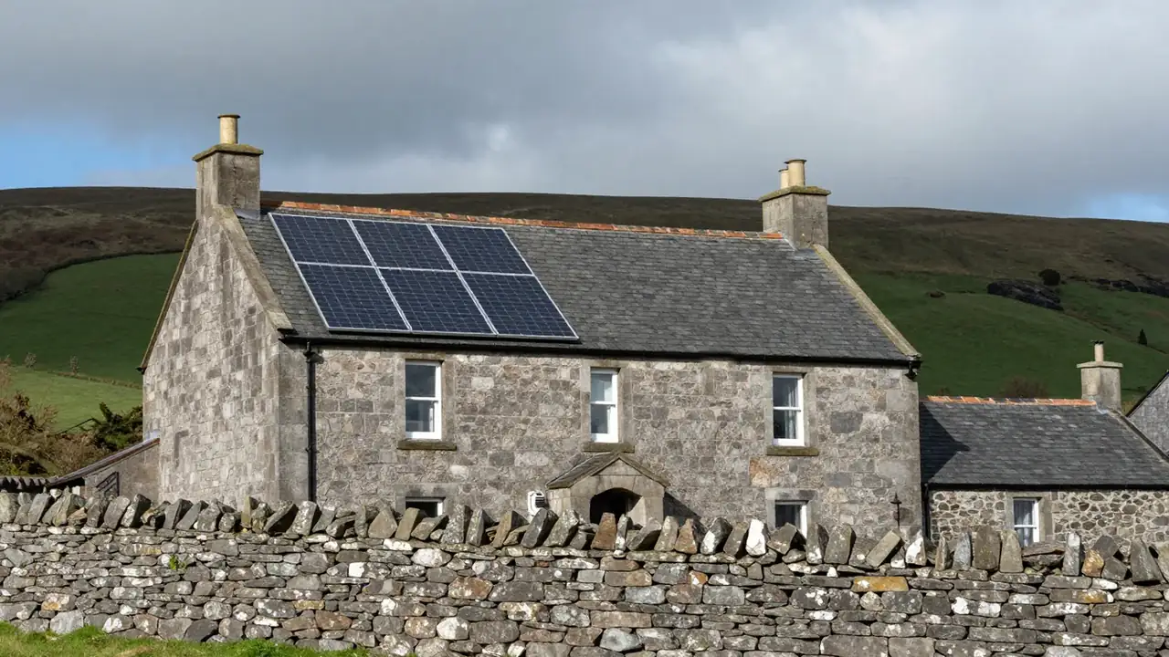 A stone-built cottage with a slate roof carrying black solar panels, behind a dry-stone wall with green hills and a cloudy sky beyond